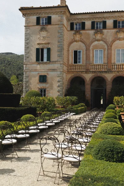 Outdoor ceremony setup with wrought iron chairs and white cushions lining a gravel aisle, framed by boxwood hedges in a villa courtyard