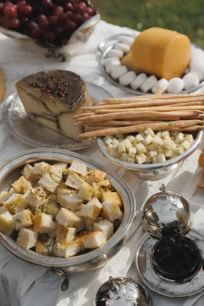 Wedding appetizer spread with cheese cubes, breadsticks, grapes and a cheese wheel on silver trays over a white tablecloth on a grass lawn
