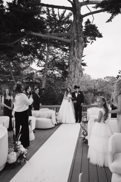 Wedding processional with a bride walking down the aisle, holding a bridal bouquet on a tree-lined wooden deck beneath open sky.