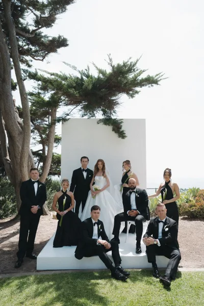 Wedding party portrait of a formal bridal party in coordinated black and white, with a calla lily bouquet, posed before trees and an ocean view.