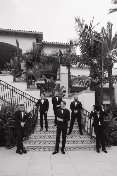 Groomsmen portrait of the groom with groomsmen in black tuxedos on a tiled staircase by a stucco building with palm trees; one wears a cowboy hat.