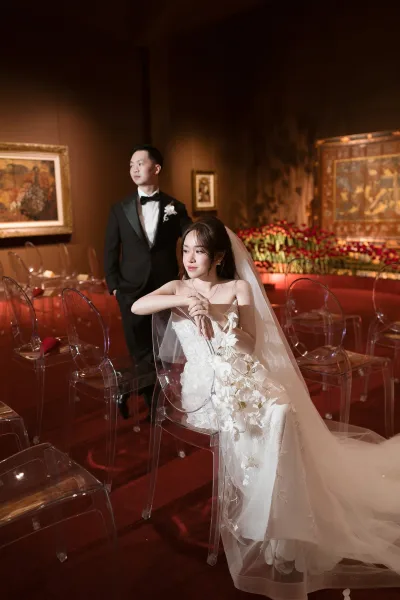 Couple portrait of bride seated in strapless lace wedding dress with cathedral veil and orchid bouquet, groom in black tuxedo on red carpet backdrop