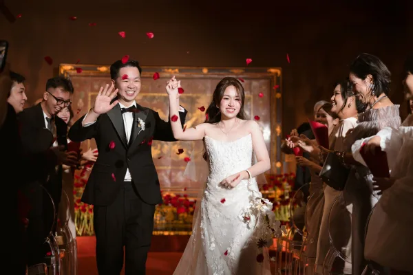 Wedding recessional as bride and groom exit holding hands while guests toss rose petals down a warm-lit indoor aisle lined with chairs