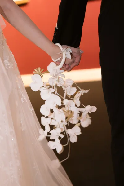Couple holding hands, bride and groom hands framed with a cascading white orchid bouquet and wrist corsage against a solid backdrop