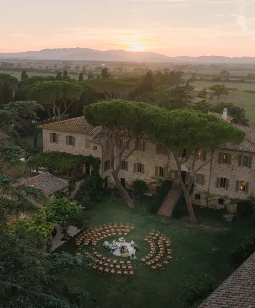 Outdoor ceremony setup with circular wedding ceremony seating around a petal-strewn aisle on a lawn by a stone villa at sunset