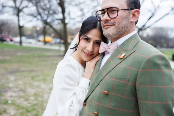 Couple portrait with the bride resting on the groom's shoulder, her long sleeve dress and a groom in a green suit on a park lawn with bare trees.