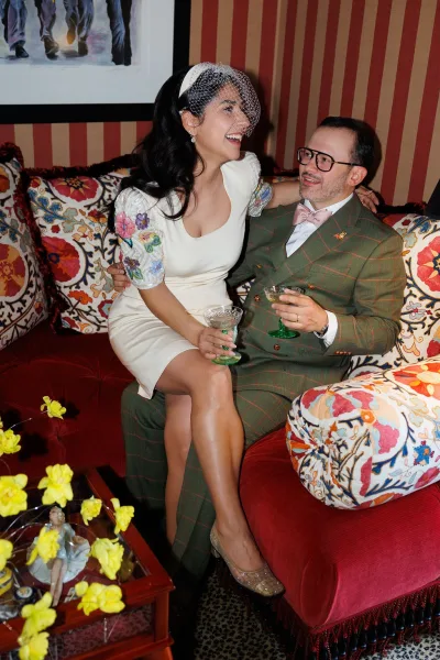 Couple portrait with a bride in a short wedding dress and groom holding coupe glasses, seated on a red velvet sofa against striped wallpaper.