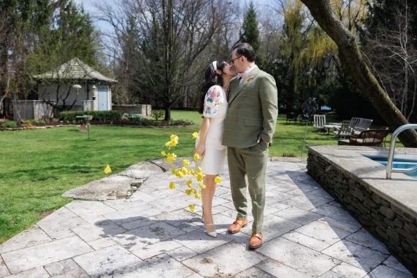 Couple portrait, a backyard wedding portrait on a stone patio by a garden gazebo, bride holding a bright yellow bouquet beside the groom.