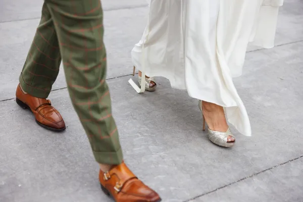 Wedding shoes laid side by side: peep toe bridal shoes with metallic heels beside the groom’s dress shoes on a concrete sidewalk.