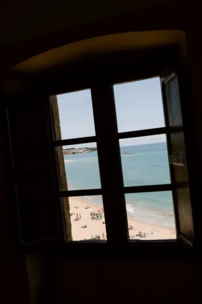 Ocean view of a sandy beach through an open window, with sailboats, a rocky islet, and distant buildings beneath a clear sky.