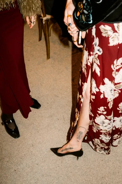 Wedding guest outfit featuring a floral guest dress and black heels, standing on gravel ground beside a wooden chair and a black tablecloth