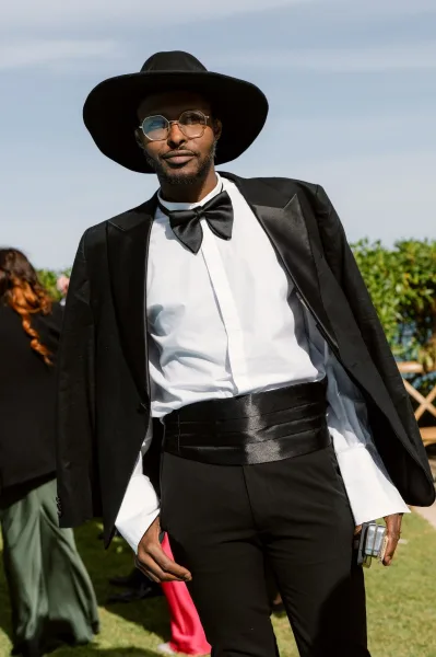 Tuxedo portrait of a man in black tie wedding attire, wearing a wide-brim hat on a grass lawn beneath a blue sky with hedges and wedding guests.