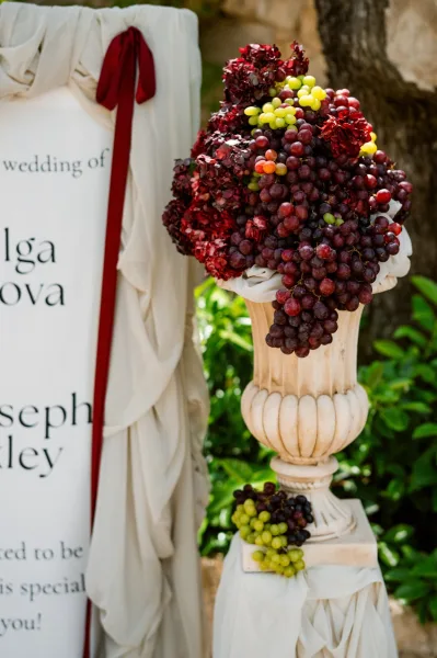 Ceremony entrance decor showcasing a wedding signage idea with cascading grapes, set against garden greenery and a weathered stone wall.