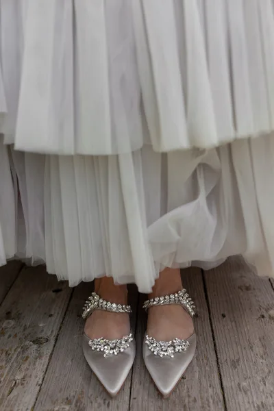 Bridal shoes with rhinestone wedding shoes sparkle, satin pointed toes and ankle straps beneath a tulle dress hem on wooden floorboards