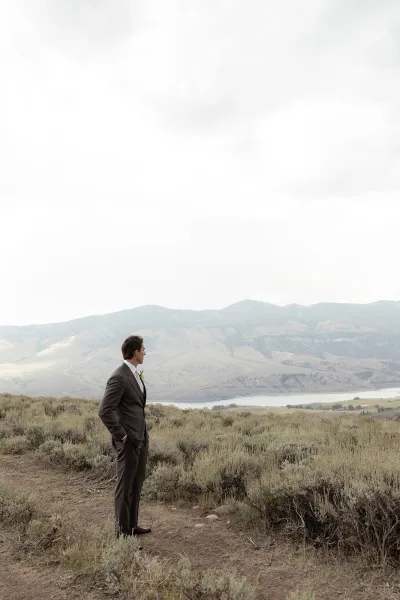 Groom portrait of a man in a brown suit with boutonniere, hands in pockets, looking away by a lake with mountains under clouds
