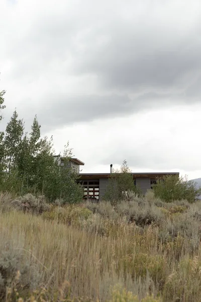 Wedding venue exterior with wood siding, large windows, and a chimney set amid tall grass field, trees, distant hills, and cloudy sky