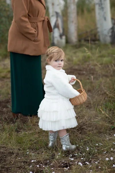 Flower girl portrait in a white tulle dress holding a wicker basket, wearing silver cowboy boots in a grassy field with petals