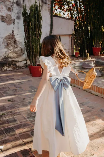 Flower girl portrait wearing a flower girl dress with a blue bow, standing on a brick patio beside a stucco courtyard fountain and potted cactus.