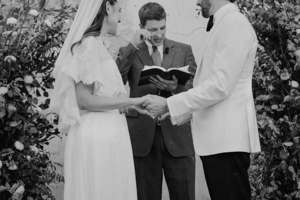 Ceremony moment as the couple holds hands during wedding vows, the bride's veil catching light before a stone wall and greenery.