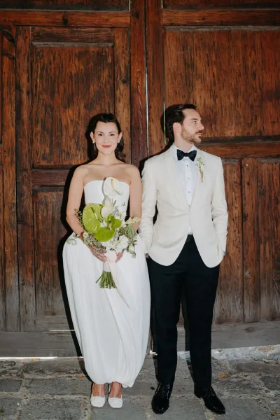 Couple portrait of a bride holding bouquet beside a groom in a white tuxedo jacket, posed against wooden doors on a stone floor.