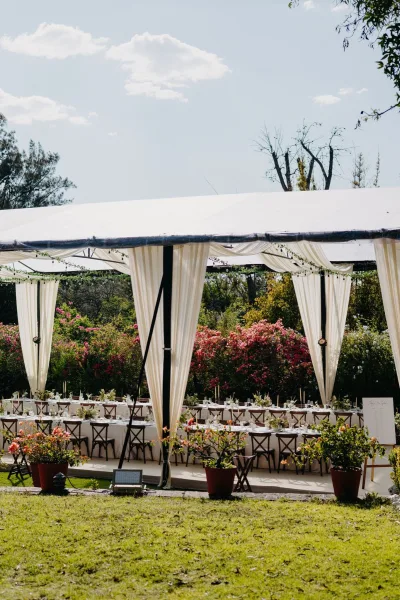 Tented reception setup showcasing refined outdoor reception decor with long banquet tables on a garden lawn beside flowering shrubs under a blue sky.
