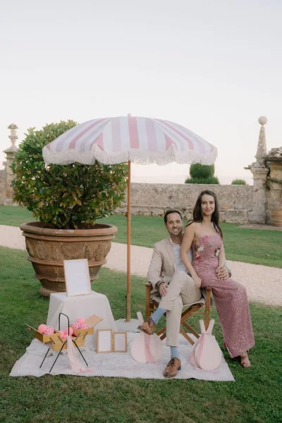 Couple portrait of a bride and groom under a striped umbrella on a garden lawn, with a gravel path and stone wall framing the scene.