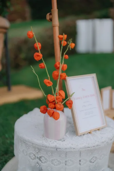 Bud vase centerpiece of Chinese lantern flowers on a lace tablecloth, set on a grass lawn with garden shrubs in the background.