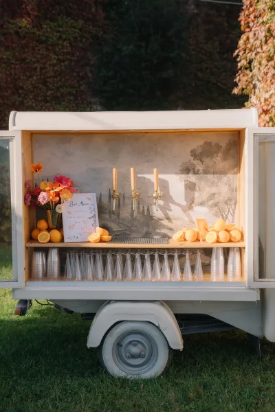 Mobile bar setup for a wedding drink station with drink taps, set on a grass lawn against an ivy-covered wall.