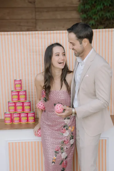Couple portrait of bride and groom laughing by a striped backdrop, her pink sequin dress gleaming as they enjoy a playful reception game moment