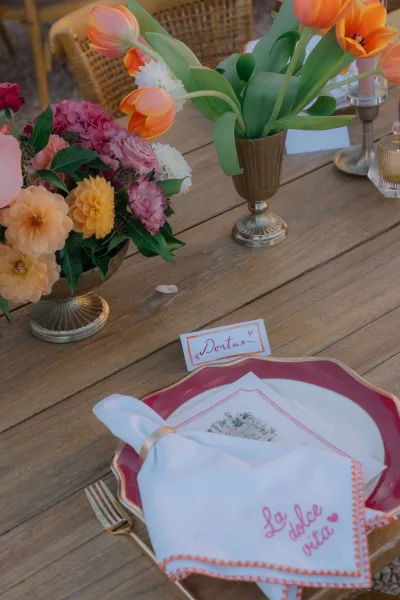 Wedding place setting styled as colorful reception table decor with a scalloped plate on a wooden table beside a rattan chair.